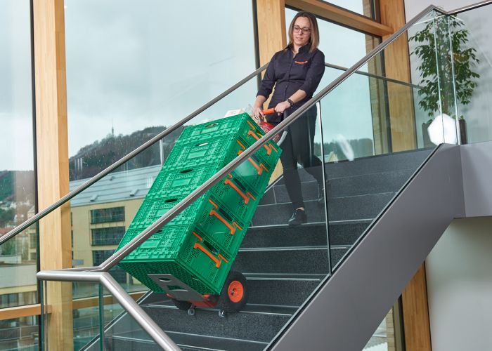 Das Bild zeigt eine Frau, die mit Hilfe eines CargoMaster Treppensteigers für Lasten mehrere Lebenmittelkisten über eine Treppe befördert. Zu sehen sind außerdem die Steigbeine des Treppensteigers sowie die Sicherheitsbremsen, die an der Treppenkante automatisch gestoppt haben.