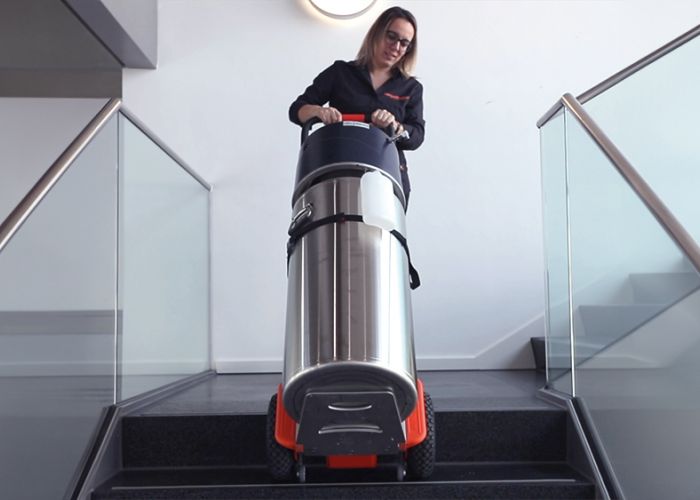 Das Bild zeigt eine Frau, die mit Hilfe eines CargoMaster Treppensteigers für Lasten einen Sauerstoffzylinder über eine Treppe befördert. Zu sehen sind außerdem die Steigbeine des Treppensteigers sowie die Sicherheitsbremsen, die an der Treppenkante automatisch gestoppt haben.