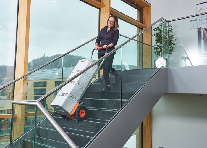 Das Bild zeigt eine Frau, die mit Hilfe eines CargoMaster Treppensteigers für Lasten mehrere Kisten Kopierpapier über eine Treppe befördert. Zu sehen sind außerdem die Steigbeine des Treppensteigers sowie die Sicherheitsbremsen, die an der Treppenkante automatisch gestoppt haben.